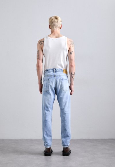 Persona rubia con tattoos en los brazos, vestida con una camiseta sin mangas blanca, jeans azul claro y zapatos oscuros, de pie frente a una pared gris lisa.