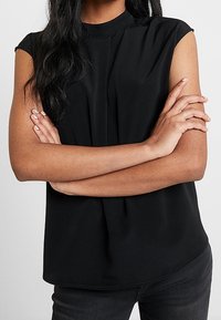 Sort kortærmet top med høj halsudskæring, diskrete plissédetaljer foran og en glat tekstur, kombineret med mørke jeans.