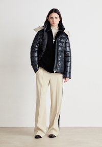Casaco puffer preto com gola bege, fecho de correr e bolsos laterais; emparelhado com calças creme com riscas laterais pretas.