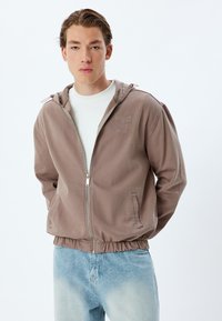 Lichtbruine zip-up jas met capuchon, elastische taille en geborduurd logo. Heeft zijzakken en een gladde, gestructureerde stof.