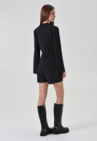 Donna con lungo capelli castani, che indossa una tuta nera a maniche lunghe e stivali neri fino al ginocchio, girata di lato su uno sfondo neutro.