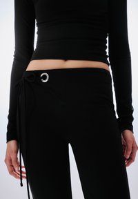 Fekete, testhez simuló nadrág sima textúrával, oldalán fém karikás megkötős záródással. Látható a hozzá illő fekete felső hosszú ujjai is.