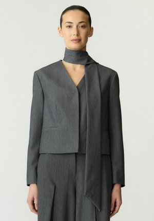 Femme portant un costume gris sur mesure avec une longue écharpe assortie drapée autour du cou, debout devant un fond clair uni.