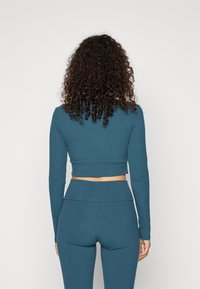 Žena s kudrnatými vlasy, oblečená v modrém žebrovaném krátkém topu s dlouhým rukávem a odpovídajících vysokých legínách, stojící na jednobarevném pozadí.