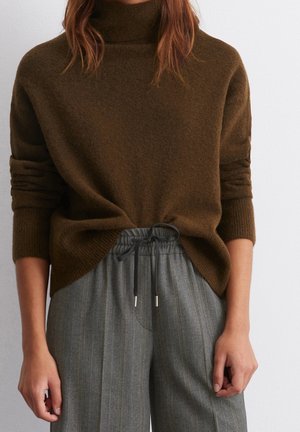 Femme portant un pull à col roulé marron et un pantalon gris à fines rayures avec cordon, debout devant un fond uni.