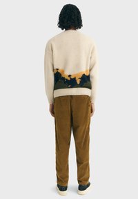 Maglione beige con un motivo astratto giallo, verde e blu navy sulla parte bassa della schiena, abbinato a pantaloni di velluto a coste marroni e sneaker nere.