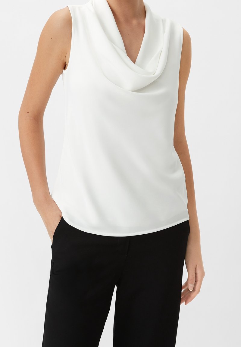 Blouse blanche sans manches avec un col drapé associée à un pantalon noir, main du modèle dans la poche sur un fond uni.