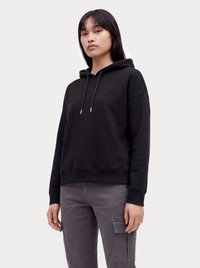 Giovane donna con felpa nera con cappuccio e pantaloni cargo grigi, in piedi davanti a uno sfondo chiaro e uniforme.