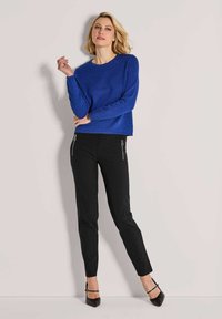Vrouw met blond haar, gekleed in een gestructureerde blauwe trui, zwarte slim fit broek met ritszakken en zwarte puntige hakken, staande tegen een lichte muur.