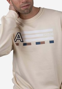 Sudadera beige con cuello redondo, que presenta un logotipo azul en forma de "A" y franjas horizontales en blanco, marrón y azul a lo largo del pecho.