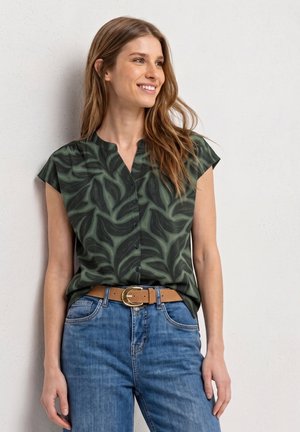 Femme souriante aux longs cheveux portant un chemisier à manches courtes motif feuilles vert foncé, ceinture marron clair et jean bleu, debout contre un mur blanc.