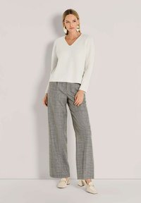 Vrouw in een witte gebreide trui, grijs geruite wijde broek, crèmekleurige blokhak instappers en geometrische gouden en witte oorbellen, poseert binnen.