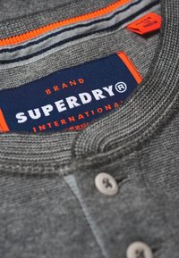 Grå strikket Superdry skjorte krave med synlige knapper, orange og marineblå mærke, samt orange størrelsesetiket indeni kraven.