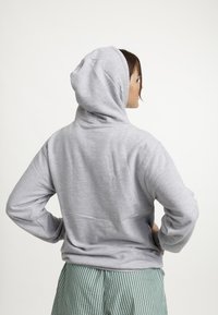 Persoon in een lichtgrijze hoodie en groen-witte gestreepte broek, zijwaarts gekeerd met handen op de heupen tegen een effen achtergrond.
