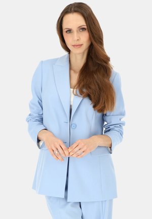 Kobieta z długimi brązowymi włosami, ubrana w jasnoniebieską marynarkę i pasujące spodnie, stojąca z rękami złożonymi przed sobą.