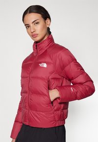 Raudona pūkinė striukė su aukštu apykakle, siuvinėtomis dalimis, užtraukiamais kišenėmis ir "North Face" logotipu ant kairiojo krūtinės. Lygus, izoliacinis audinys.