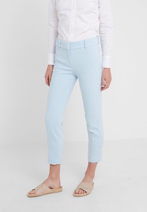 Personne portant un pantalon slim bleu clair aux chevilles, une chemise blanche boutonnée et des sandales beige à enfiler, debout devant un fond blanc uni.