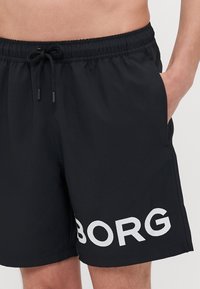 Sungas pretas com uma cintura elástica, fecho de cordão, bolsos laterais e letras brancas perfuradas "BORG" na perna esquerda.