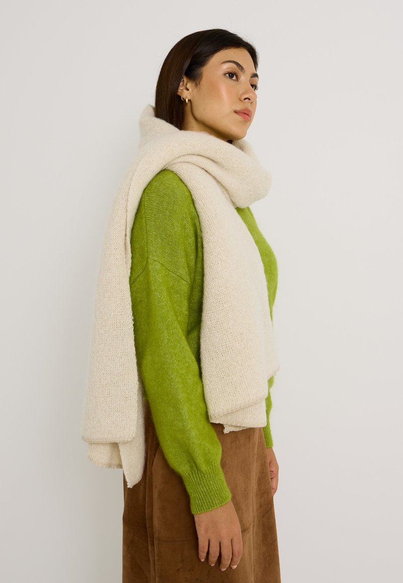 Bufanda beige texturizada colocada sobre un suéter verde y una falda marrón, mostrando materiales suaves y colores contrastantes.