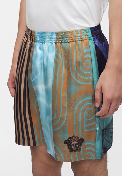 Homme portant un short coloré à motifs avec des dessins abstraits bleus, orange et noirs et un emblème tête de Méduse près de l'ourlet.