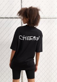 Juoda per didelė t-shirt marškiniai su užrašu "CHEERS!" baltos rašytinės tekstūros ant nugaros. Modelis derina juodus dviračių šortus.