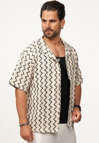 Kurzarmhemd in Creme mit einem schwarzen Zickzackmuster, ausgestattet mit einem Kragen, Knopfleiste und lockerer Passform. Über einem schwarzen Tanktop getragen.