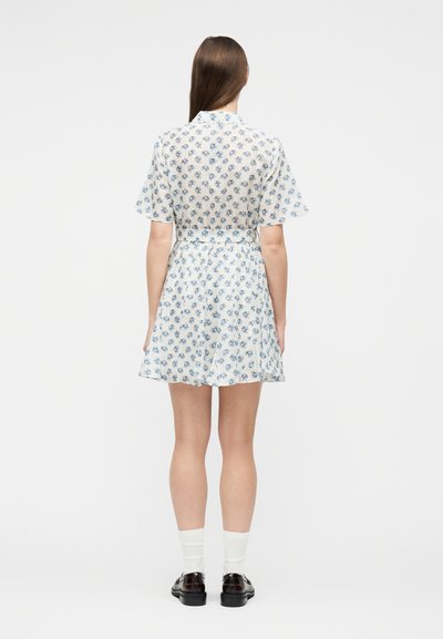 Femme debout, dos tourné, portant une mini-robe blanche à manches courtes avec un imprimé floral bleu, des chaussettes blanches et des mocassins noirs.