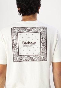 Bílý bavlněný tričko s velkým hnědým grafickým motivem lemovaným paisley na zadní straně a textem "Barbour Outdoor Clothing" ve středu.