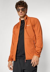 Oransje zip-up jakke med teksturert stoff, svarte detaljer og to sidelommer. Brukt over en svart skjorte, kombinert med runde solbriller.