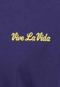 Camiseta morada con texto amarillo "Vive La Vida". La tela tiene una textura suave y acanalada con un diseño ajustado.