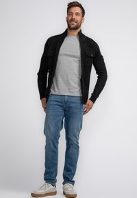 Zwarte gebreide cardigan met een rits, twee borstzakken; gecombineerd met een grijs T-shirt, lichtblauwe jeans en grijze sneakers met bruine zolen.