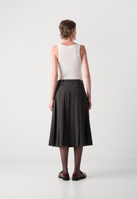Personne portant un haut sans manches blanc, une jupe midi plissée gris foncé, des collants noirs et des mocassins marron, debout face à un mur clair.