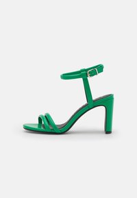 Sandale en cuir verni vert dotée d'un talon fin, d'une lanière réglable à la cheville et de trois lanières parallèles sur les orteils. Texture lisse.
