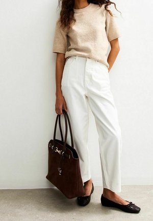 Femme portant un pull beige à manches courtes, un pantalon blanc taille haute, des ballerines noires, tenant un grand sac à main marron foncé, debout contre un mur blanc.