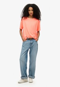Camiseta de manga corta de color rosa neón, de corte relajado, combinada con jeans rectos de color azul claro y zapatillas blancas. Un simple cinturón marrón acentúa el conjunto.