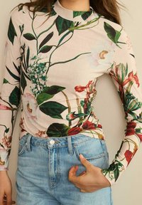 Long-sleeve top in light pink fabric with a floral pattern featuring green leaves, red and pink flowers. Paired with light blue high-waisted jeans.