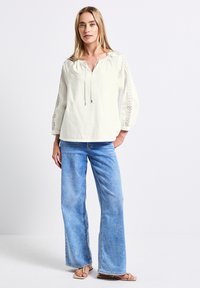 Vit långärmad blus med spetser detaljer på ärmarna, matchad med ljusblå vida jeans och sandaler. Texturerat bomullstyg.