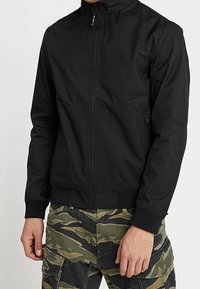 Svart lättviktsjacka med hög krage, hel dragkedja fram och elastiska ärmslut, kombinerad med kamouflagefärgade cargobyxor.