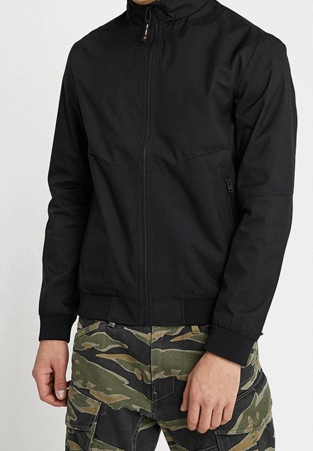Svart lättviktsjacka med hög krage, hel dragkedja fram och elastiska ärmslut, kombinerad med kamouflagefärgade cargobyxor.