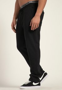 Svarta sweatpants med elastisk midja, grå märkning, sidofickor och avslappnad passform, kombinerat med svarta sneakers.