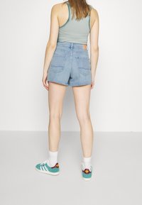 Shorts de denim, de color azul claro, de cintura alta con bolsillos traseros. Llevados con una camiseta sin mangas de color verde claro y zapatillas en tono aguamarina con acentos rojos.