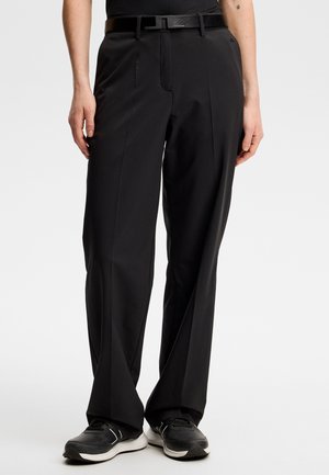 Osoba nosząca czarne spodnie o szerokich nogawkach z czarnym paskiem oraz czarno-białe trampki, stojąca na jednolitym tle.