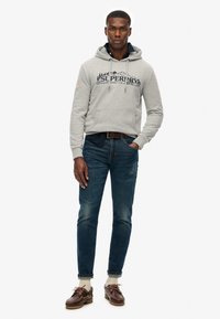 Grijze hoodie met een voorzak en een marineblauw logo, gecombineerd met blauwe spijkerbroeken, bruine veterschoenen en crèmekleurige sokken. Model staat tegen een witte achtergrond.