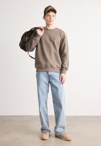 Brun genser, lyseblå baggy jeans og beige sko. Komplett med en grønn caps med beige detaljer og en mørkebrun sekk.