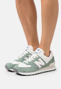 Gröna och vita New Balance 574 sneakers, tillverkade av mocka och läder, med en strukturerad sula, logotypdetaljer och platta skosnören.