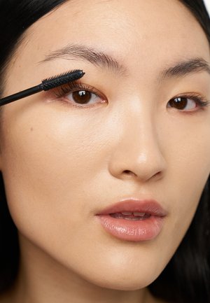 Zwarte mascara wordt aangebracht op de wimpers met een speldraadarige borstel. De huidtint is gelijkmatig, met een natuurlijke, lichte glans op de lippen.