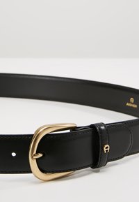 Cinturón de cuero negro con una hebilla de metal dorado, que presenta una textura suave y un detalle de logo bordado. Diseño clásico con acentos simples.