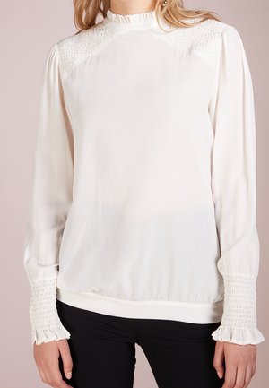 Vrouw die een witte blouse met lange mouwen draagt, voorzien van geplooide schouders, een gerimpelde hoge kraag en manchetten, gecombineerd met een zwarte broek tegen een effen achtergrond.