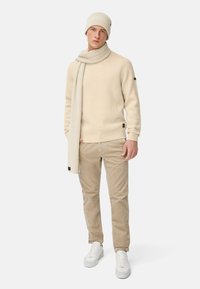 Beiger gestrickter Pullover, passender Schal, hellbeige Cord-Hose und weiße Sneakers. Gerippte Textur, dezente Logo-Details an den Ärmeln.