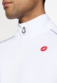 Weißer Radfahrmantel mit hohem Kragen, vorderem Reißverschluss und kleinem roten Logo. Aus glattem Material und mit grauen Akzenten auf den Schultern.